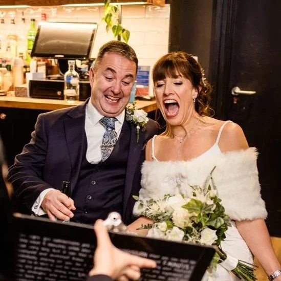 A bride and groom, dressed in wedding attire, are smiling and reacting with joy as they see their live caricature portrait revealed during their wedding reception.