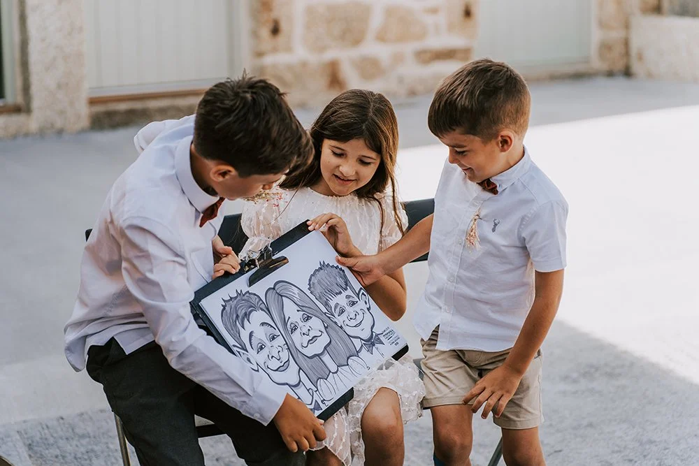 Two boys and a girl looking at a caricature drawing of three smiling faces on paper, outdoors at a wedding.