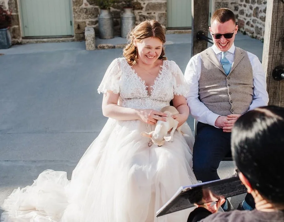 Woman in a wedding dress holding a small dog, sitting beside a man in a vest and sunglasses, smiling during a wedding celebration outdoors.