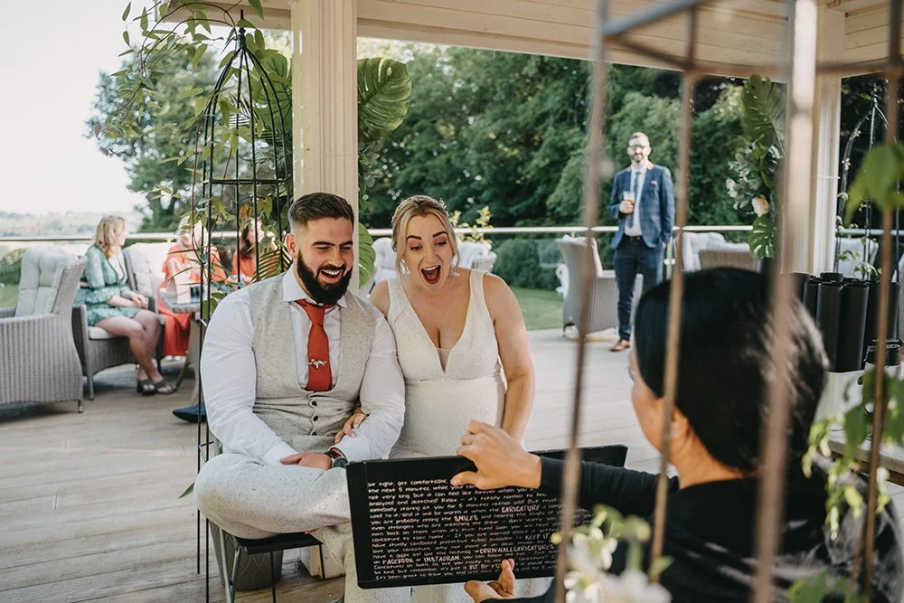 A newly married couple, a man and a woman, sitting outdoors at a wedding reception, looking at a caricature on a board with joy and laughter, with guests and greenery in the background.