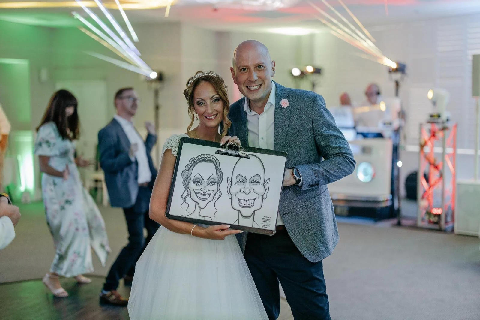 A couple at a wedding holding a framed caricature drawing of themselves, smiling and posing together in a decorated event space with other guests and colorful lighting in the background.