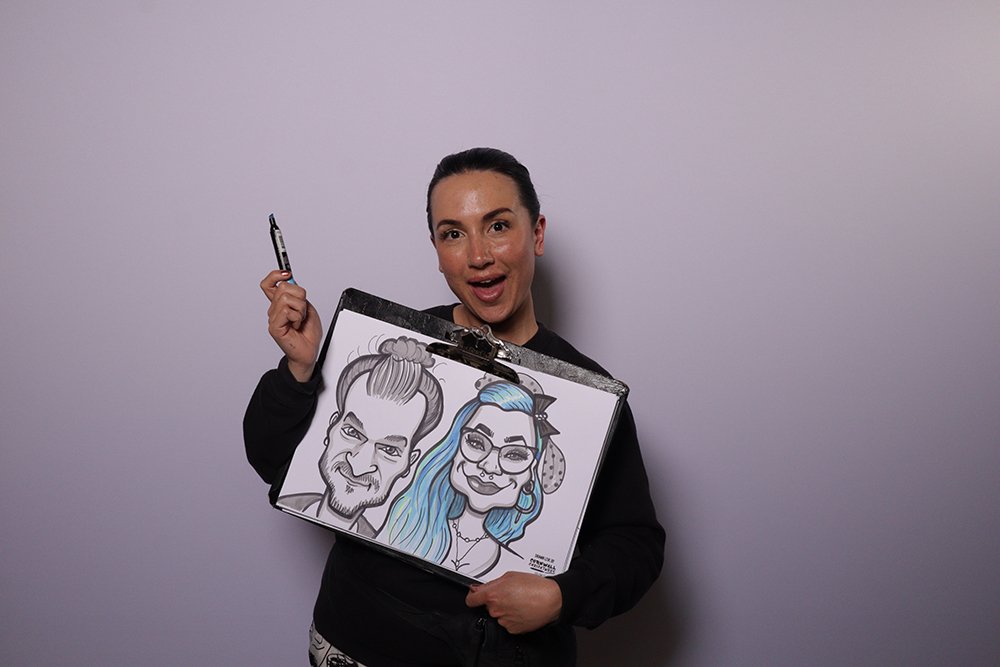Woman holding a drawing of two caricature portraits, one of a man and one of a woman, with a pen in her other hand, standing against a plain background.