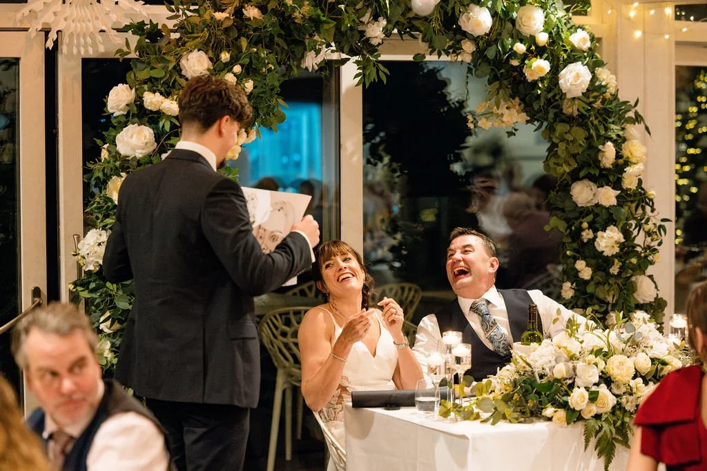 A wedding reception with a bride and groom sitting at a table, laughing as a man in a suit holds a caricature. The table is decorated with white flowers and candles, and a floral arch is overhead.