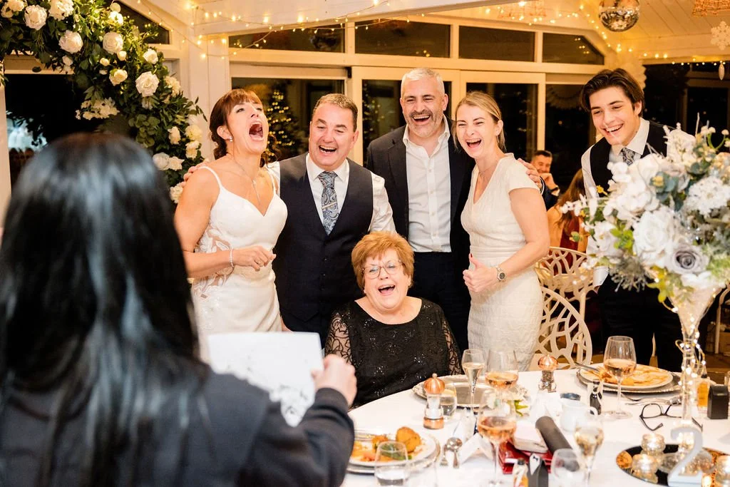 Group of six people smiling and laughing at a wedding reception, seated and standing around a table with food and drinks, decorated with flowers, in an elegant venue with string lights.