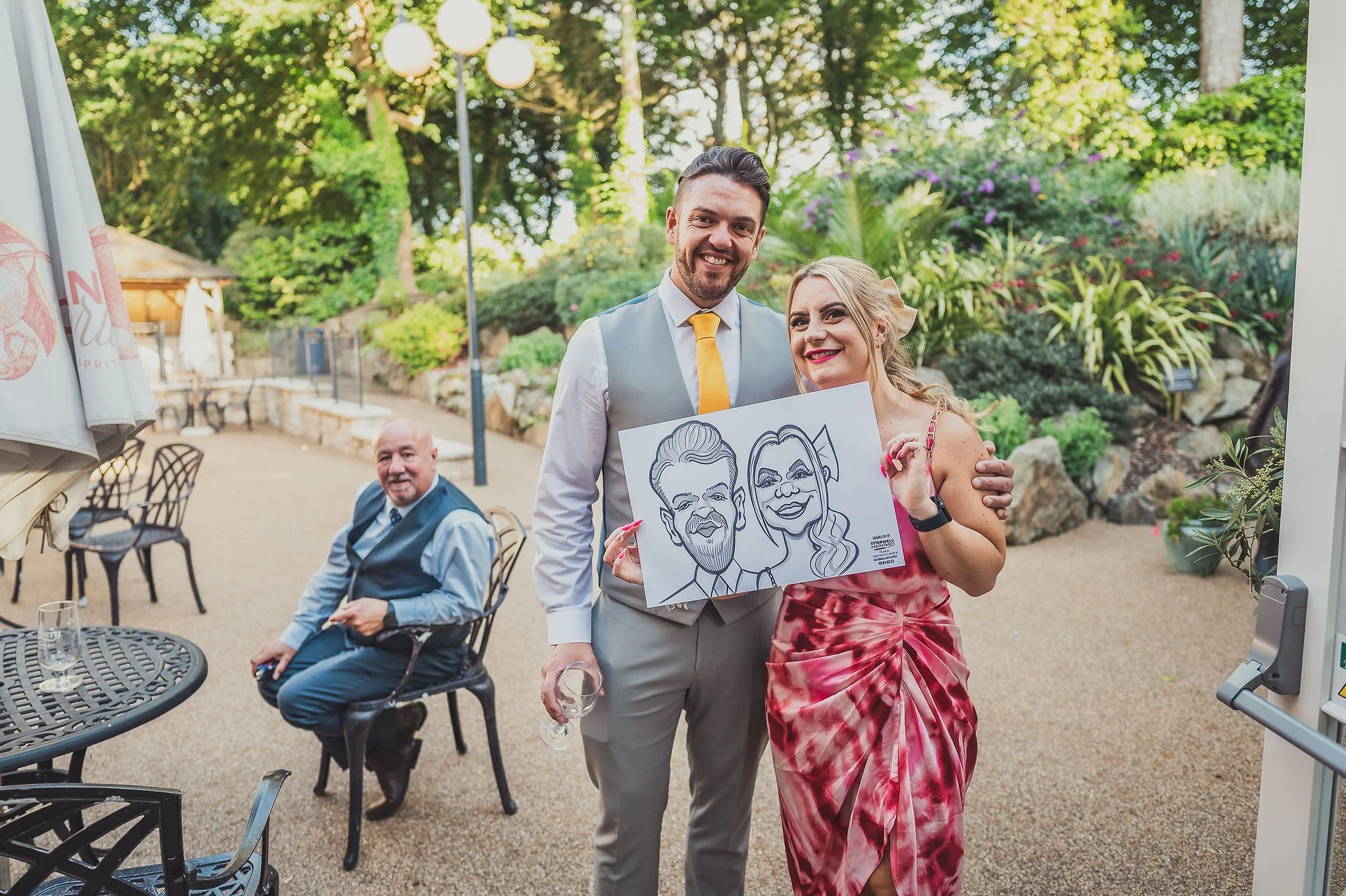A group of people at an outdoor event, with a woman holding a caricature portrait of herself and a man next to her smiling. There are tables, chairs, and lush greenery in the background.