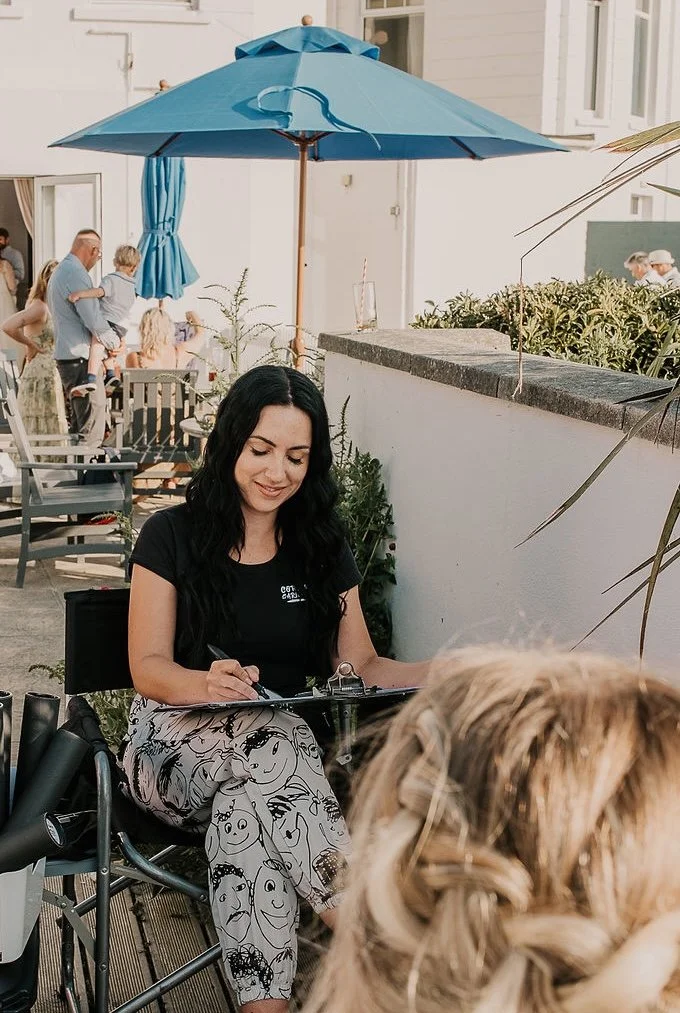 A woman with long black hair sitting outdoors at a table, smiling and drawing, with a large blue umbrella overhead, in a lively patio setting.