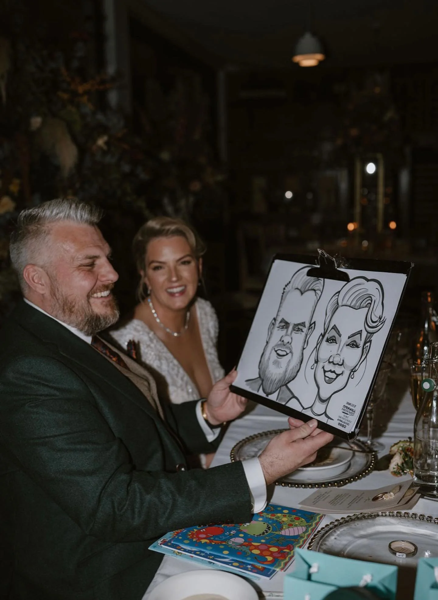 A man and woman at a table during a celebration, with the man holding a caricature drawing of himself and the woman, seated in front of a decoration-covered wall.