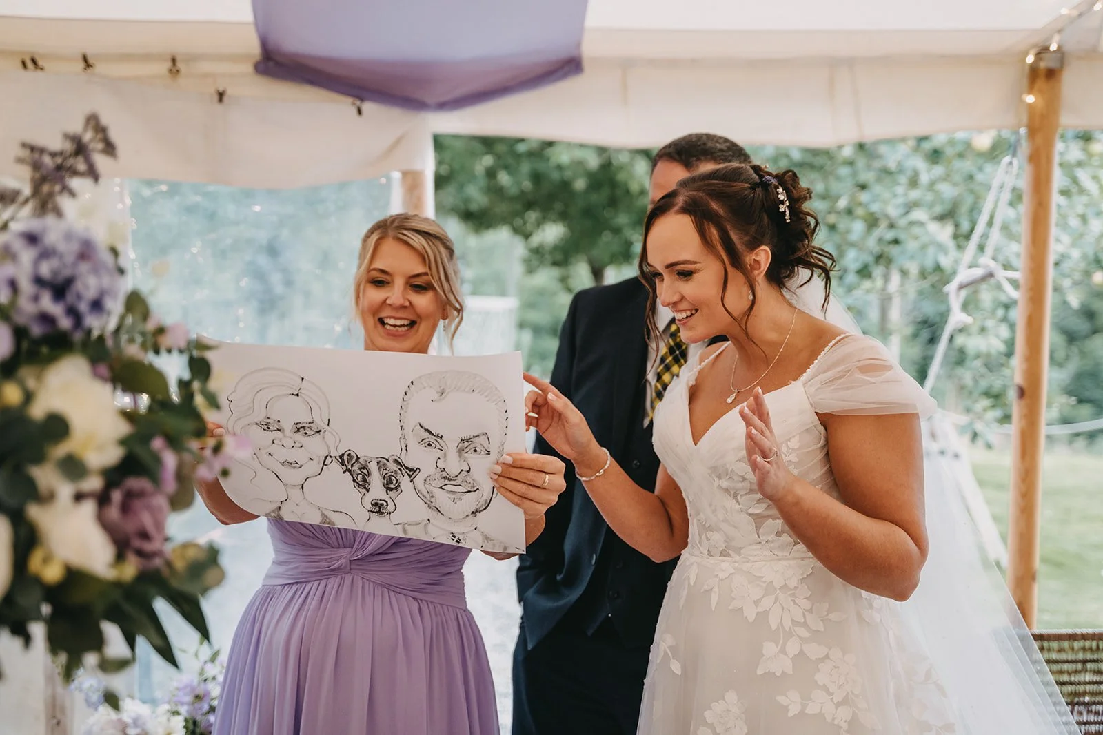 A bride, wearing a white lace wedding dress, is looking at a caricature drawing of herself, her partner, and a dog, held by a woman in a lavender dress at a wedding reception outdoors.