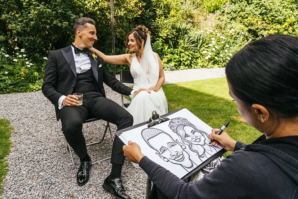 A bride and groom sitting outdoors, smiling and looking at each other, while a caricature artist draws their portraits. The bride is in a white dress with a veil, and the groom is in a tuxedo. The caricature artist is capturing their faces on a sketc