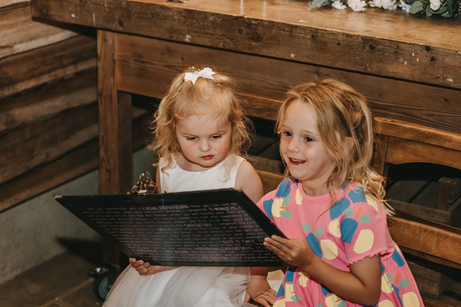 Two young girls sitting on a wooden bench, looking at a caricature with interest. One girl with curly blonde hair in a white dress, and the other girl with wavy light brown hair in a colorful dress.