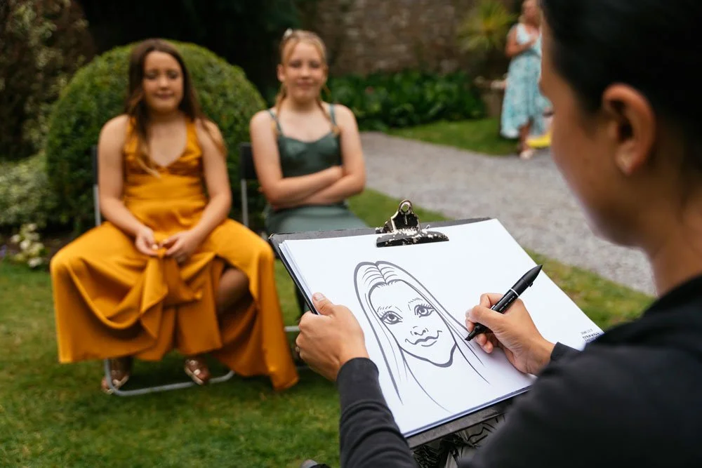 An artist sketching two young girls sitting outdoors on chairs, with a woman standing in the background.