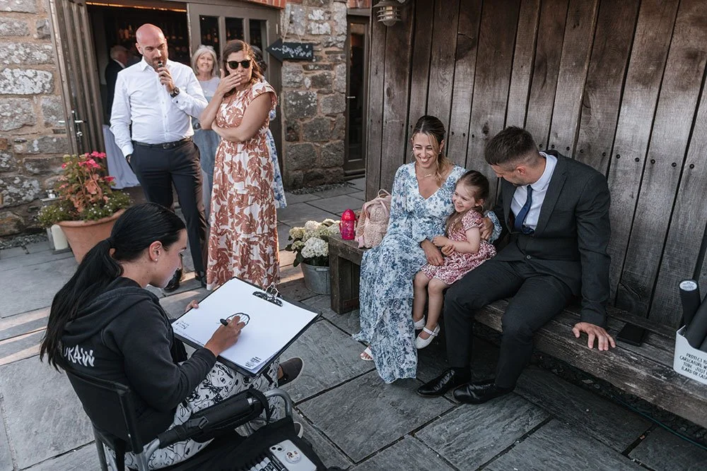 A woman in a floral dress and a young girl are sitting on a wooden bench with a man in a suit in an outdoor setting, smiling and looking at a woman drawing a caricature. Several other people are standing nearby, observing.
