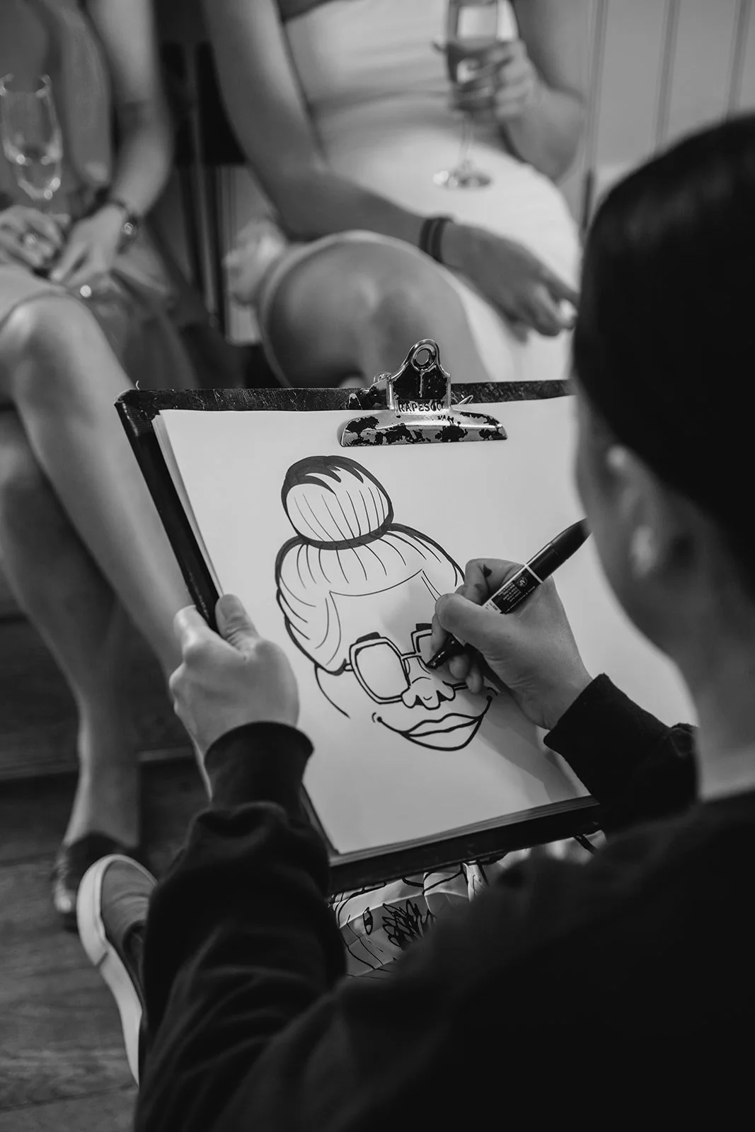 An artist drawing a cartoon portrait of a woman with her hair in a bun and wearing sunglasses on a clipboard. Two women are seated in the background, one holding a glass of wine and the other with crossed legs.
