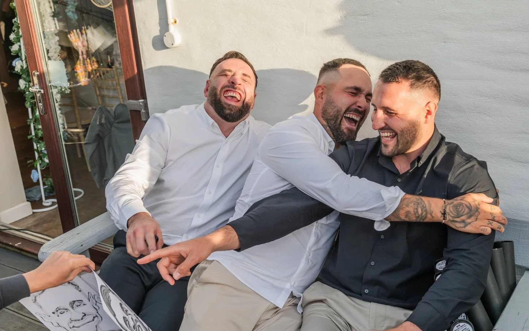 Three men laughing and hugging outside on a sunny day, sitting on a bench near a white wall with a sliding door and decorated interior visible behind.