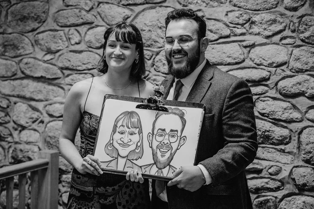 A man and a woman standing together, smiling, holding a framed caricature of themselves, in front of a stone wall.