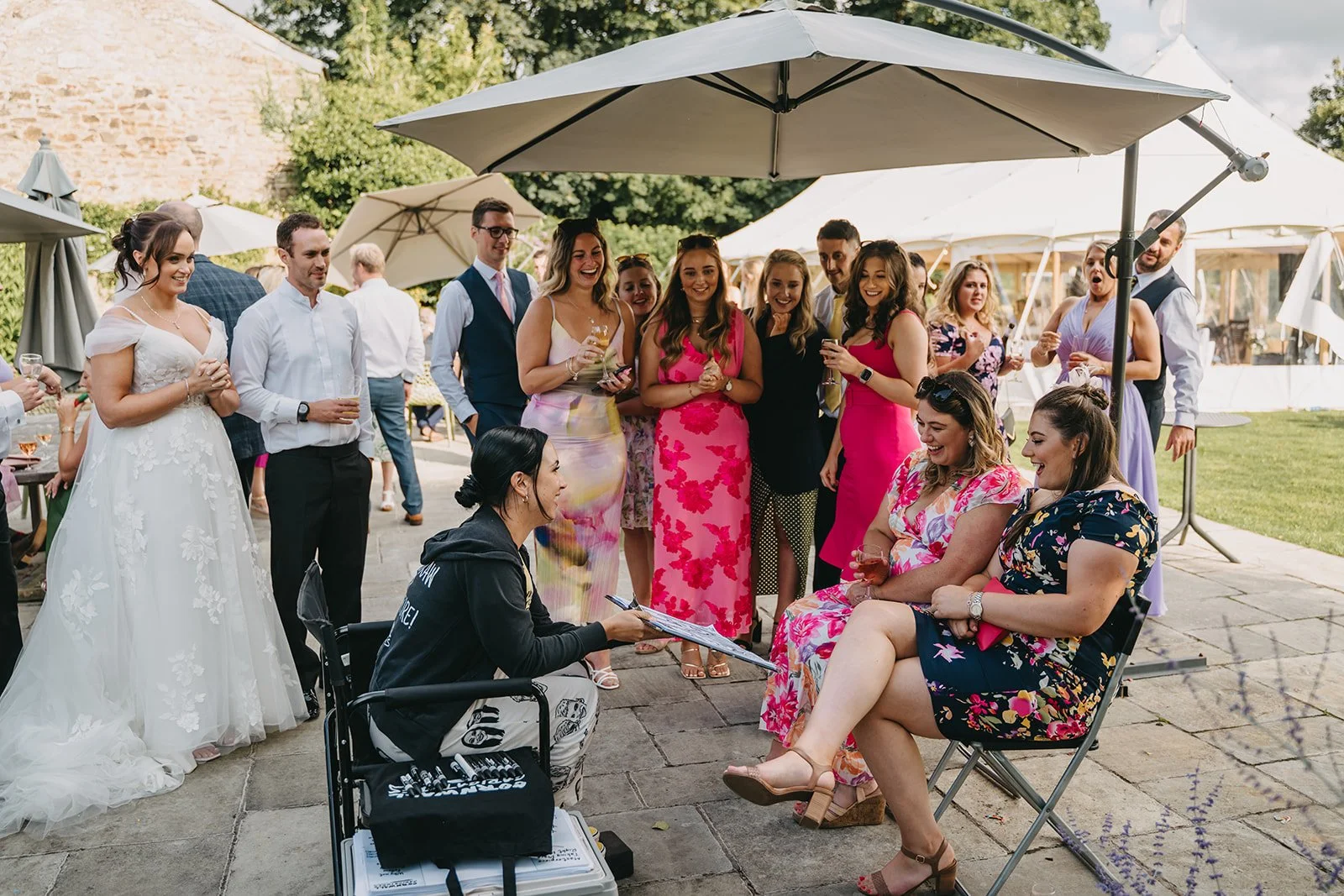 A group of people gathered outdoors at a wedding reception, with some sitting getting caricatures drawn and others standing, smiling and talking, under large umbrellas.