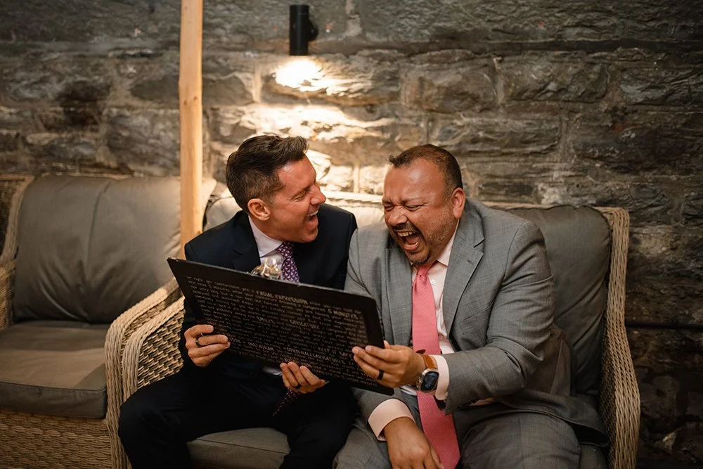 Two men in suits laughing and sharing a caricature at a wedding with a stone wall background.