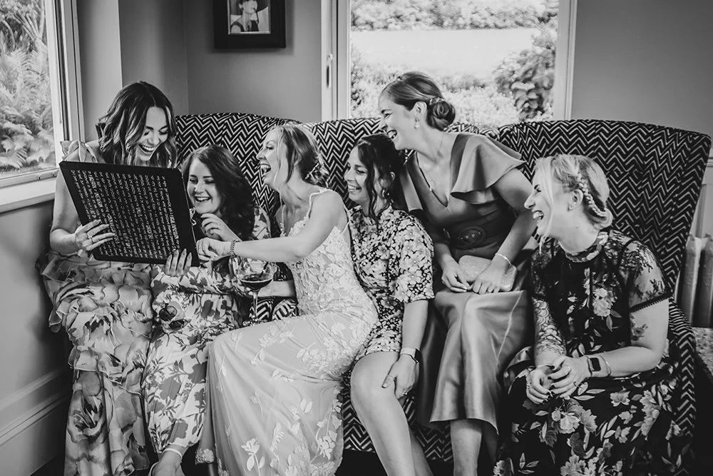 Six women, including a bride in a wedding dress, sitting on a couch, smiling and laughing while looking at caricature artwork.