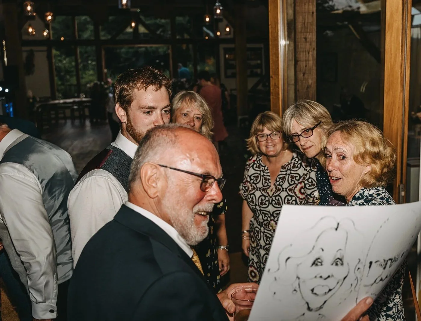 Group of adults, mostly women, gathered around an older man with glasses, looking at a drawing of a face on a large piece of paper at a social event in a rustic venue.
