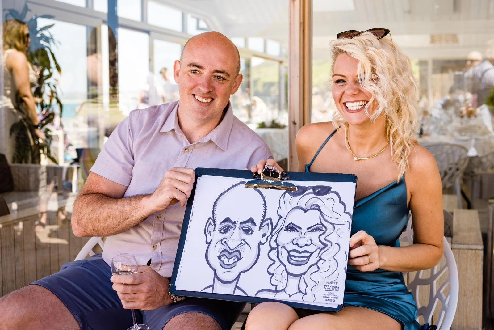 A man and a woman sitting at a table in a bright, sunny restaurant, holding a caricature drawing of themselves on a clipboard. The man is bald, wearing a light pink shirt, and the woman has curly blonde hair, wearing a blue dress. They are smiling an