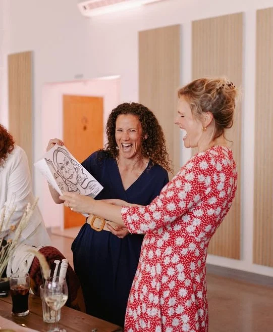 Two women laughing and looking at a drawing, at a social gathering or celebration, with drinks on the table.