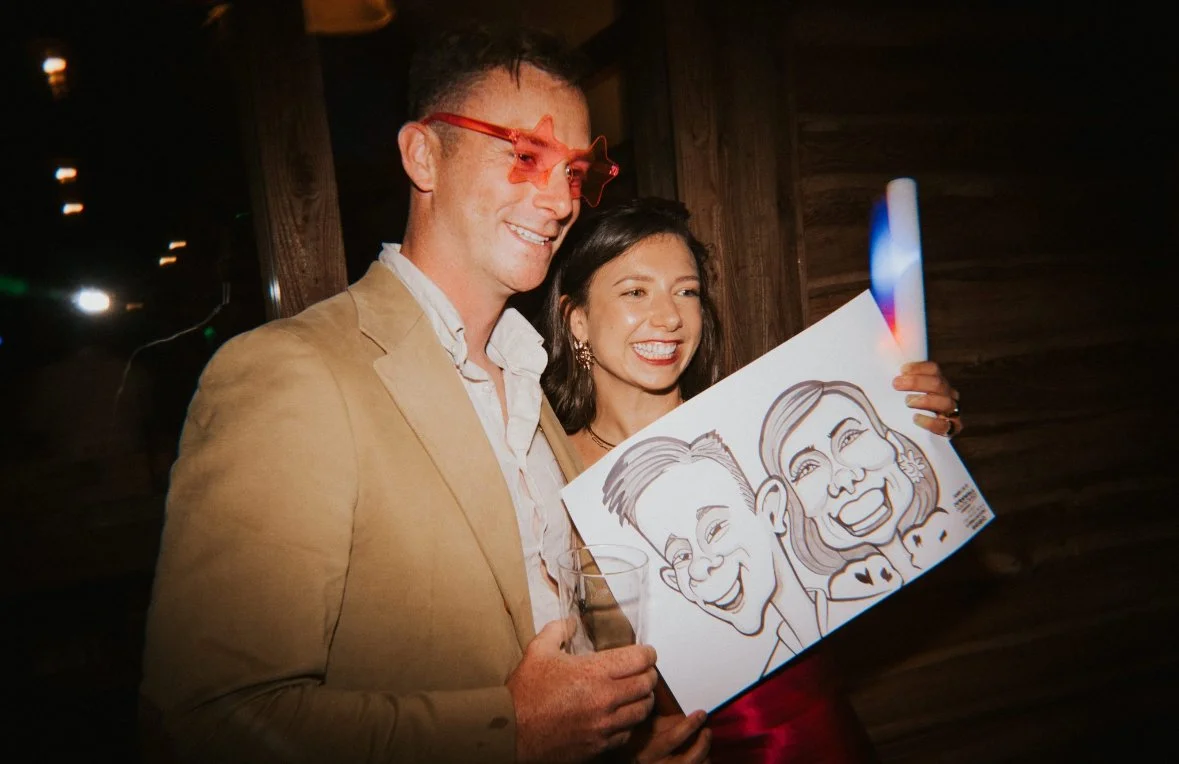 A man and woman smiling and holding a caricature drawing of themselves at a party or celebration.