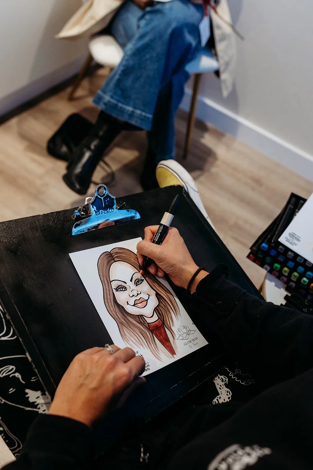 Person drawing a caricature of a woman with long hair at a desk, another person sitting in the background.