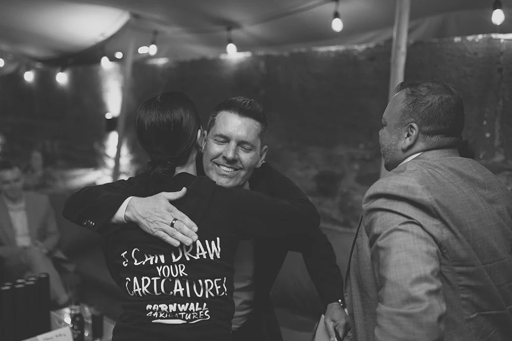 People hugging at a social event under string lights, in a tented outdoor setting, black and white photograph.