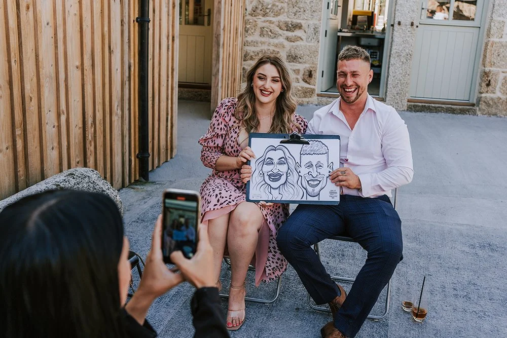 Two people sitting outdoors, smiling and holding a caricature drawing of themselves, while another person takes a photo of them.