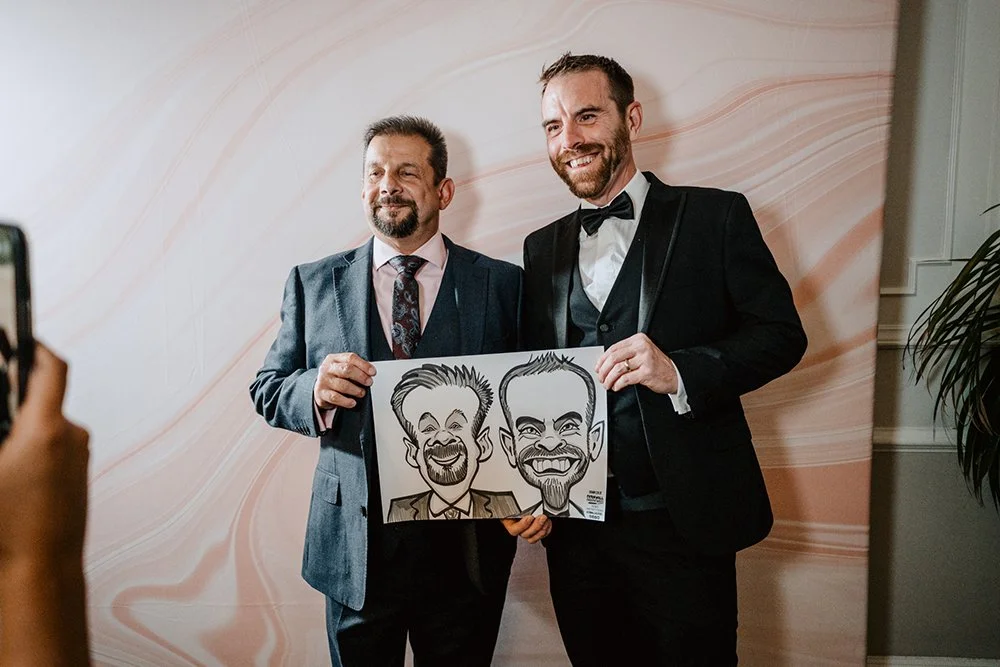Two men in suits holding a caricature drawing of themselves, standing in front of a pink marbled wall at a formal event.