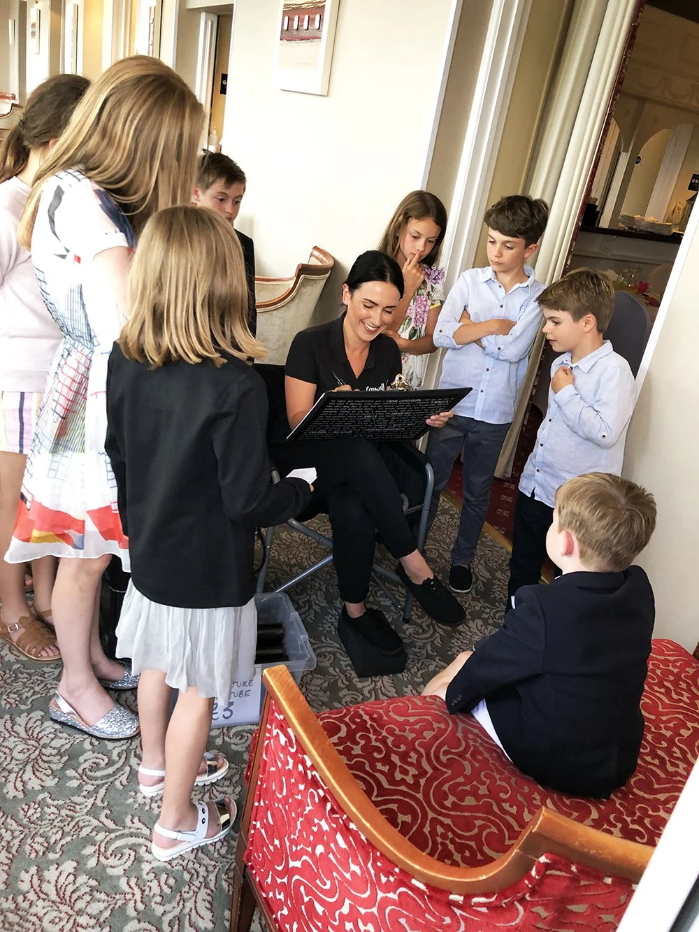 A woman sitting on a chair surrounded by children, drawing a caricature, while the children stand around her, some leaning in and others observing.