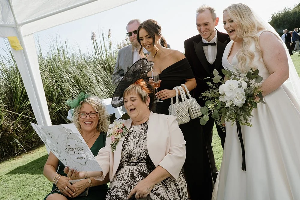 Group of people at a wedding celebration outdoors, including a bride holding a bouquet, women wearing hats and dresses, and men in suits. Some are laughing and enjoying the moment.