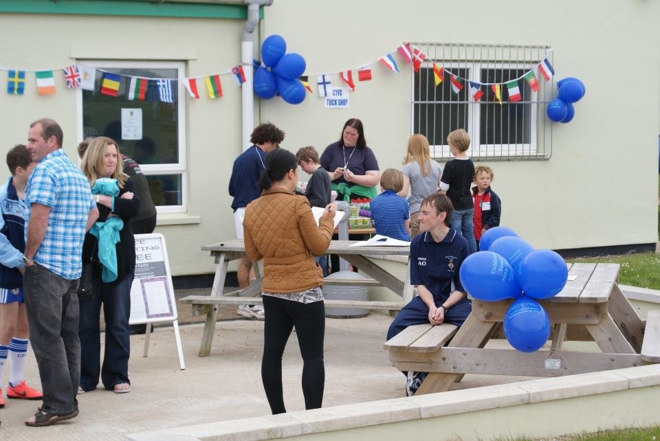 Children and adults gathered outside a building decorated with international flags and blue balloons for a community or school event.