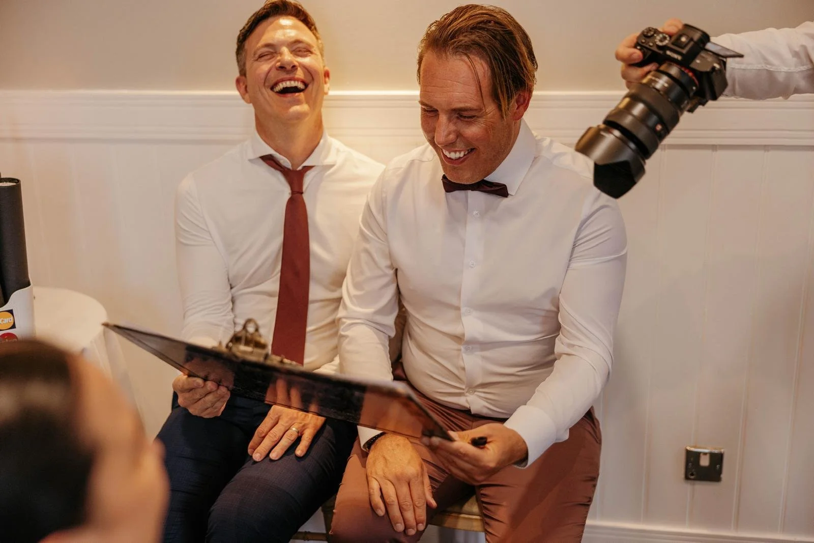 Two men dressed in white shirts and bow ties sitting together, smiling and laughing while looking at a clipboard. One man has a camera being held by another person partially visible in the picture.