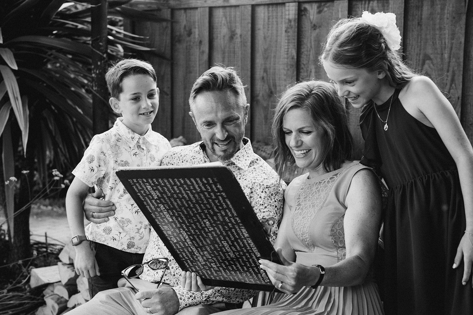 Black and white photo of a family of four gathered outdoors, looking at a large caricature, with the father and mother sitting and the two children standing beside them, all smiling and engaged.