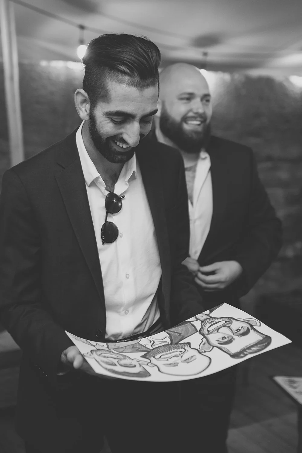 Two men in suits, smiling, one holding a caricature drawing of themselves in a casual setting.