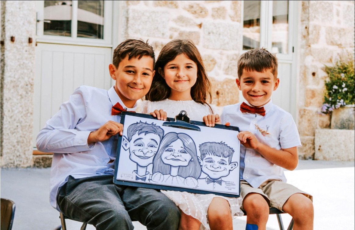 Three children, two boys and a girl, sitting outdoors with a caricature drawing of themselves, smiling at the camera.