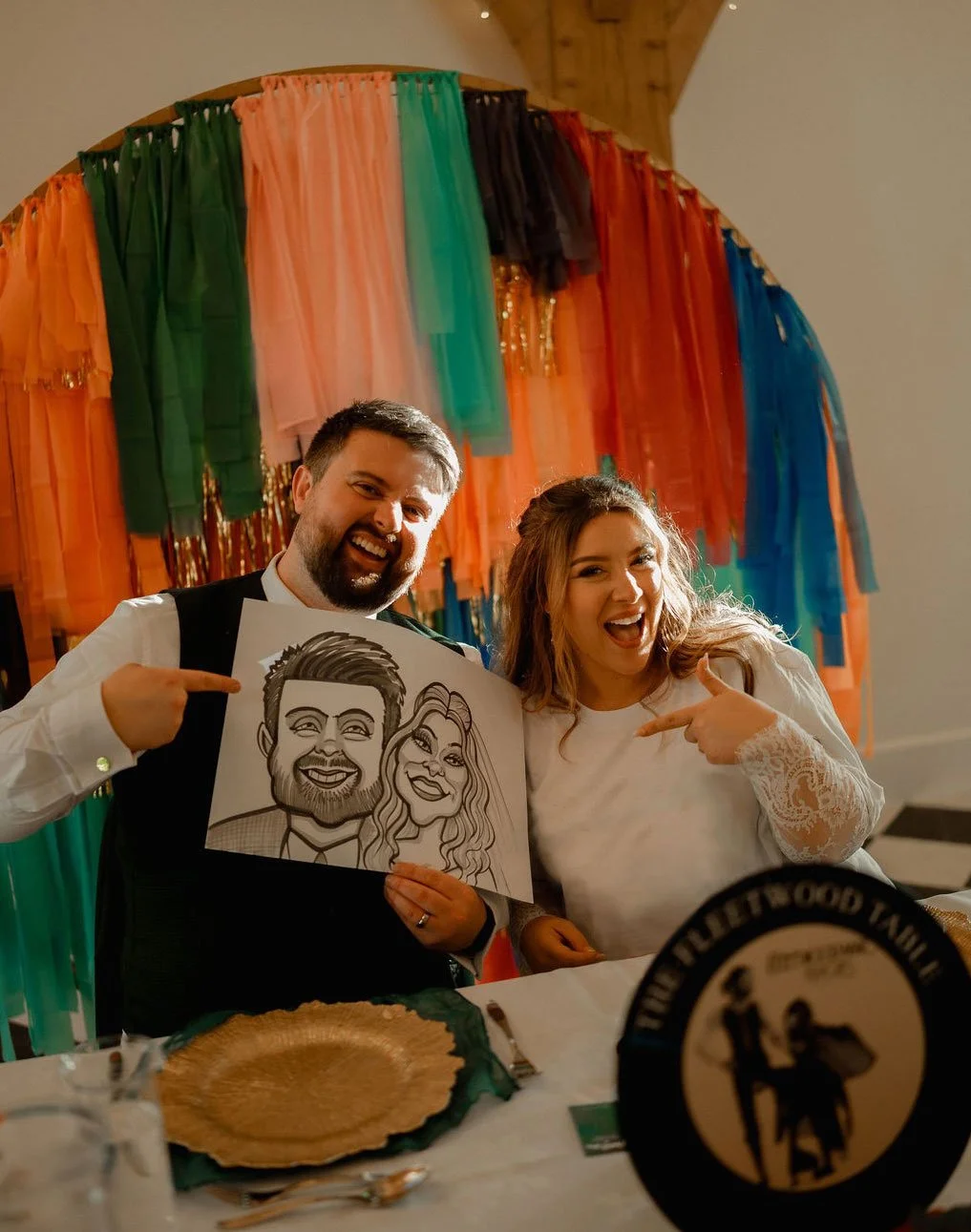 A joyful man and woman posing at their wedding, holding a caricature drawing of themselves, with a colourful rainbow fabric backdrop behind them.