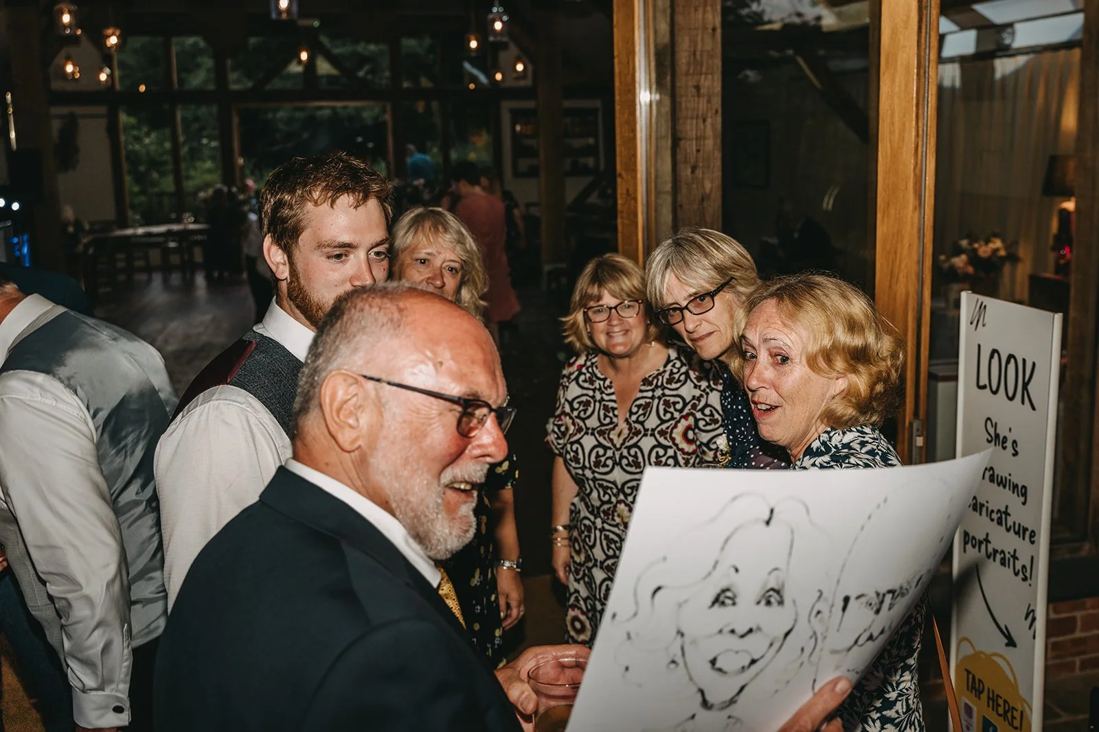 Group of people gathered indoors, looking at a drawing or caricature of a woman's face on paper.
