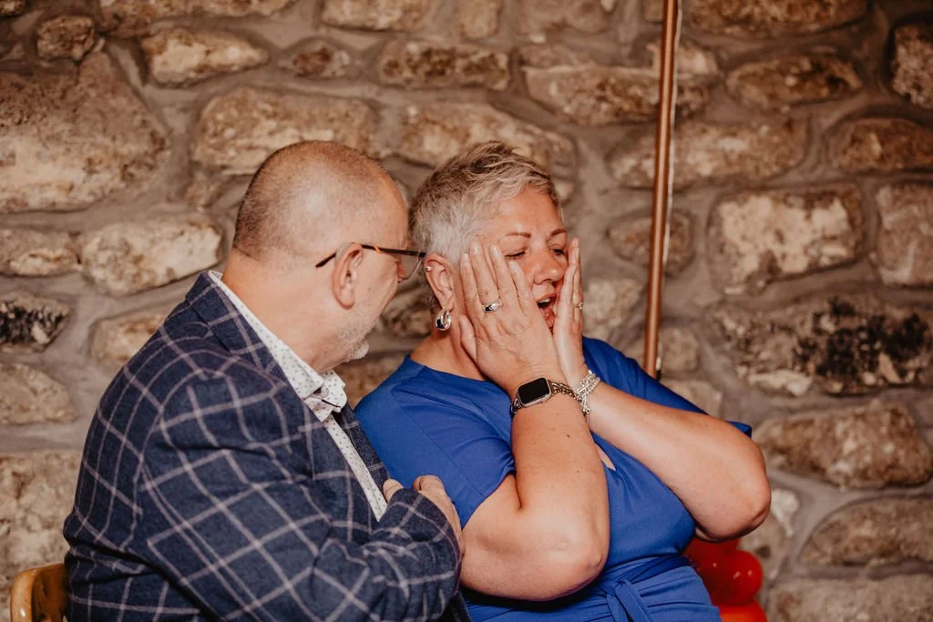 A man and a woman are sitting close, with the woman showing a surprised or happy reaction. The man is looking at her, and she has her hands on her cheeks. The background features a rustic stone wall.