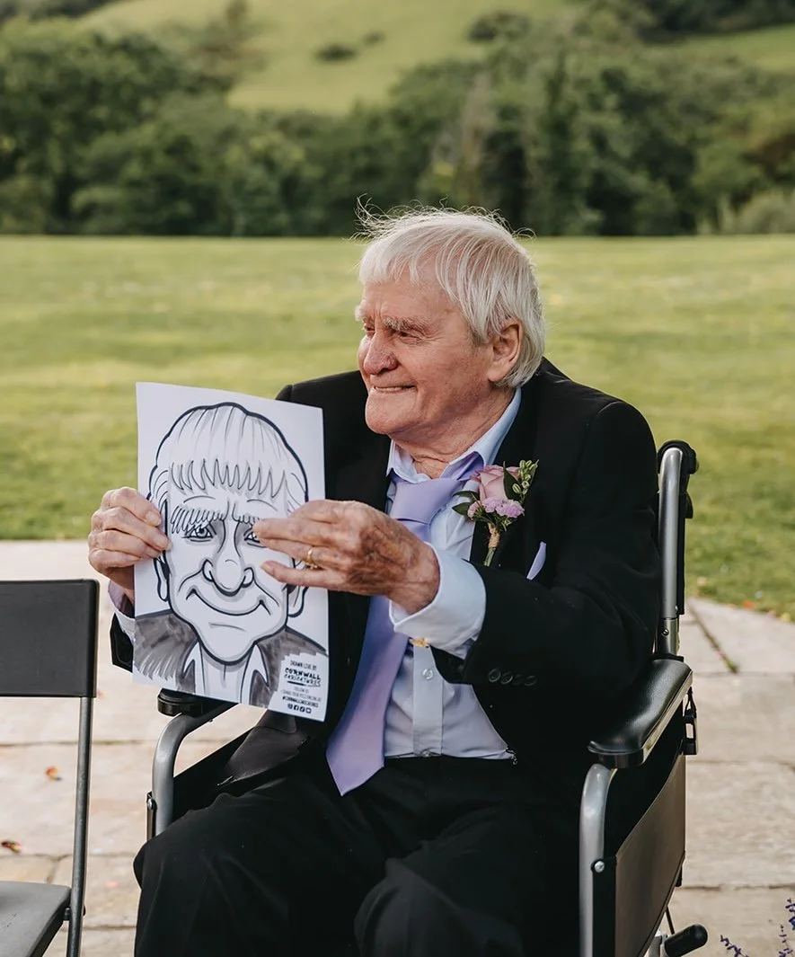 An elderly man in a wheelchair wearing a black suit and light purple tie holding a caricature portrait of himself with a smiling expression outdoors on a grassy area.