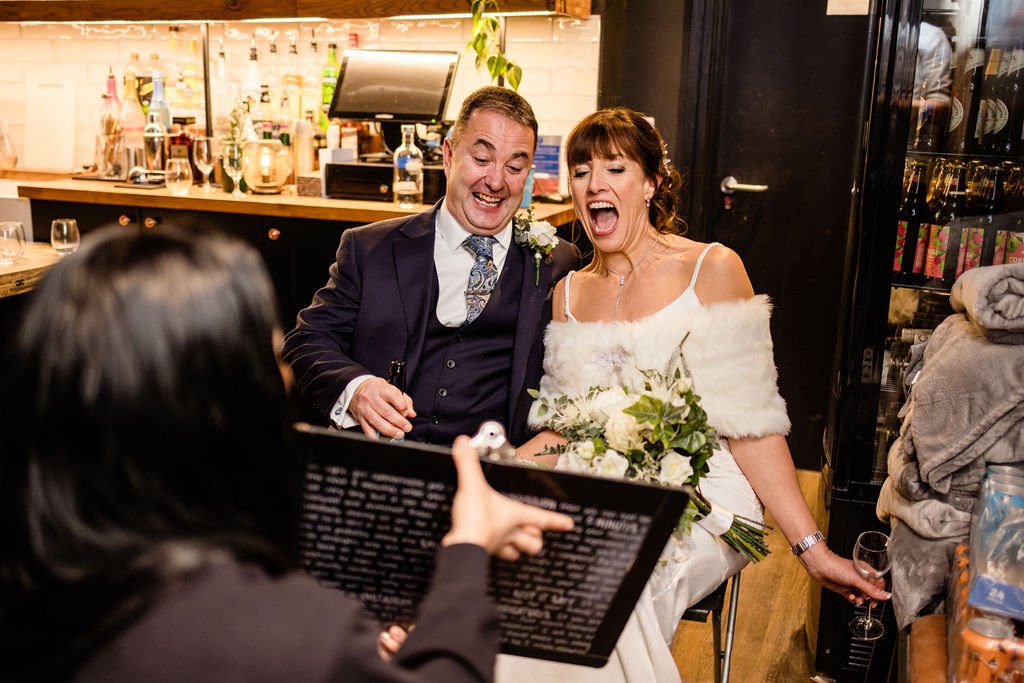 A bride and groom sitting together at their wedding reception, smiling and laughing  The bride wears a white dress with a fur shawl and holds a bouquet, and the groom wears a suit with a boutonniere. A person in the foreground is holding a caricature