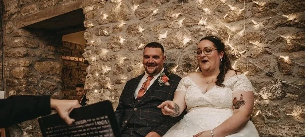 A bride and groom sit together during their wedding to get their caricatures drawn. They are indoors with a rustic stone wall decorated with string lights behind them.