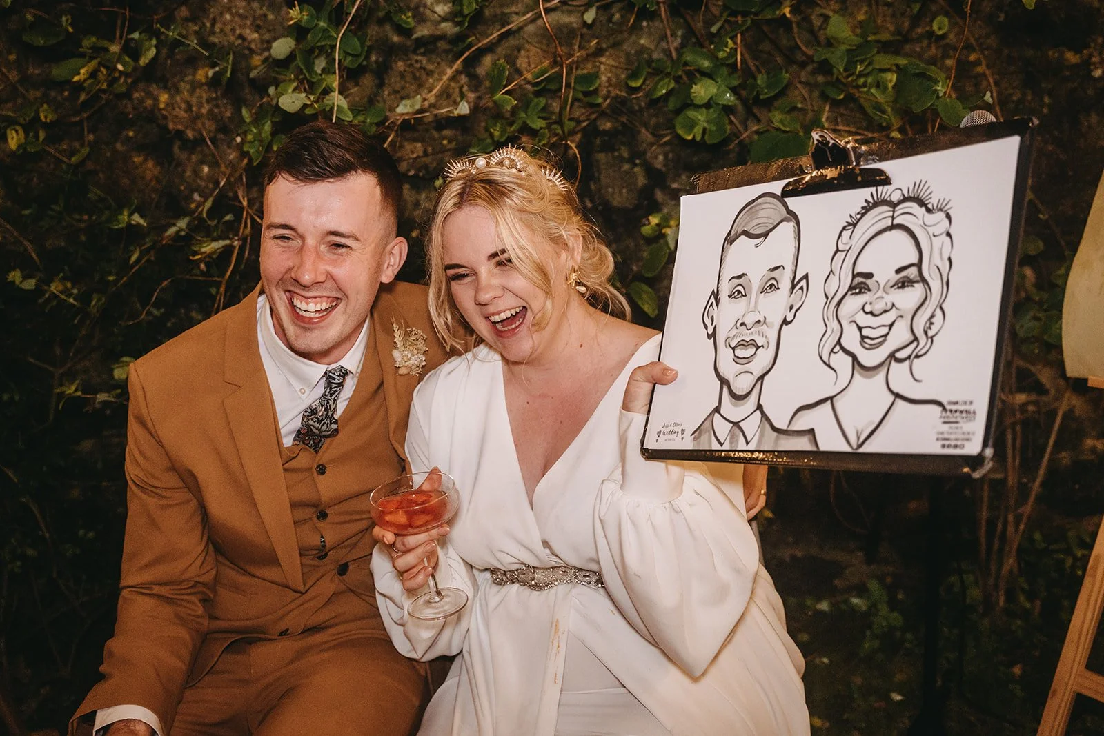 A smiling man and woman at a celebration, sitting beside each other. The woman is holding a drink, and the man is wearing a brown suit. The woman is holding a caricature artwork of a man and woman, both smiling, on a clipboard.
