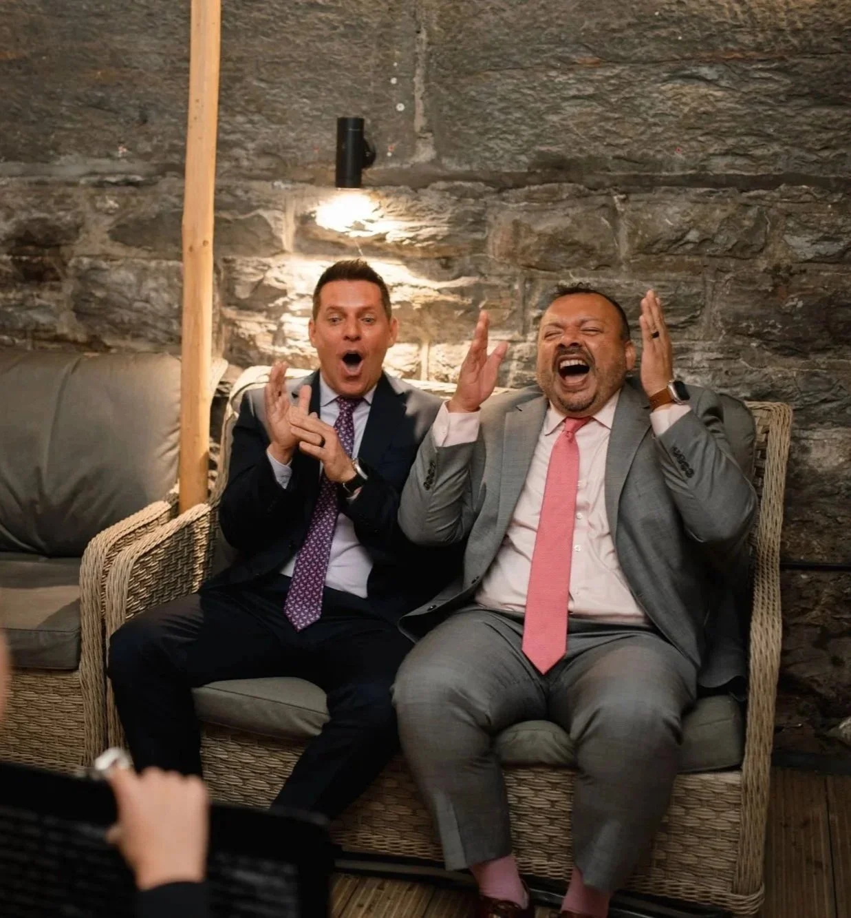 Two men in suits sit on a wicker sofa against a stone wall, appearing surprised or excited to see their caricature drawn live, with open mouths and hands near their heads.