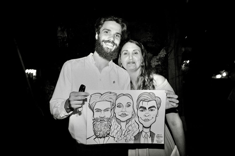 A man and woman in formal attire smiling and holding a caricature drawing of themselves against a dark background.