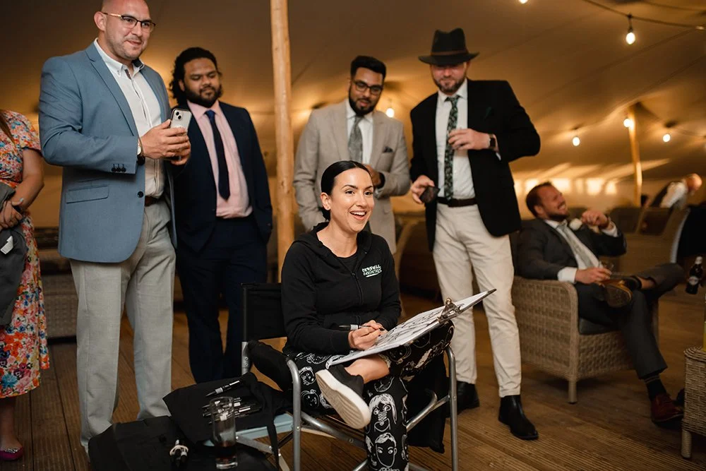 A group of people at an indoor event, with a woman sitting in a chair holding a clipboard or sketchpad, surrounded by men in suits and casual attire, some holding drinks and one taking a photo.