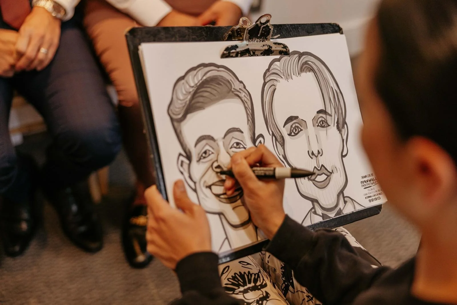 A person drawing a caricature of two individuals on a paper pad using a black marker, with the caricature featuring exaggerated facial features.