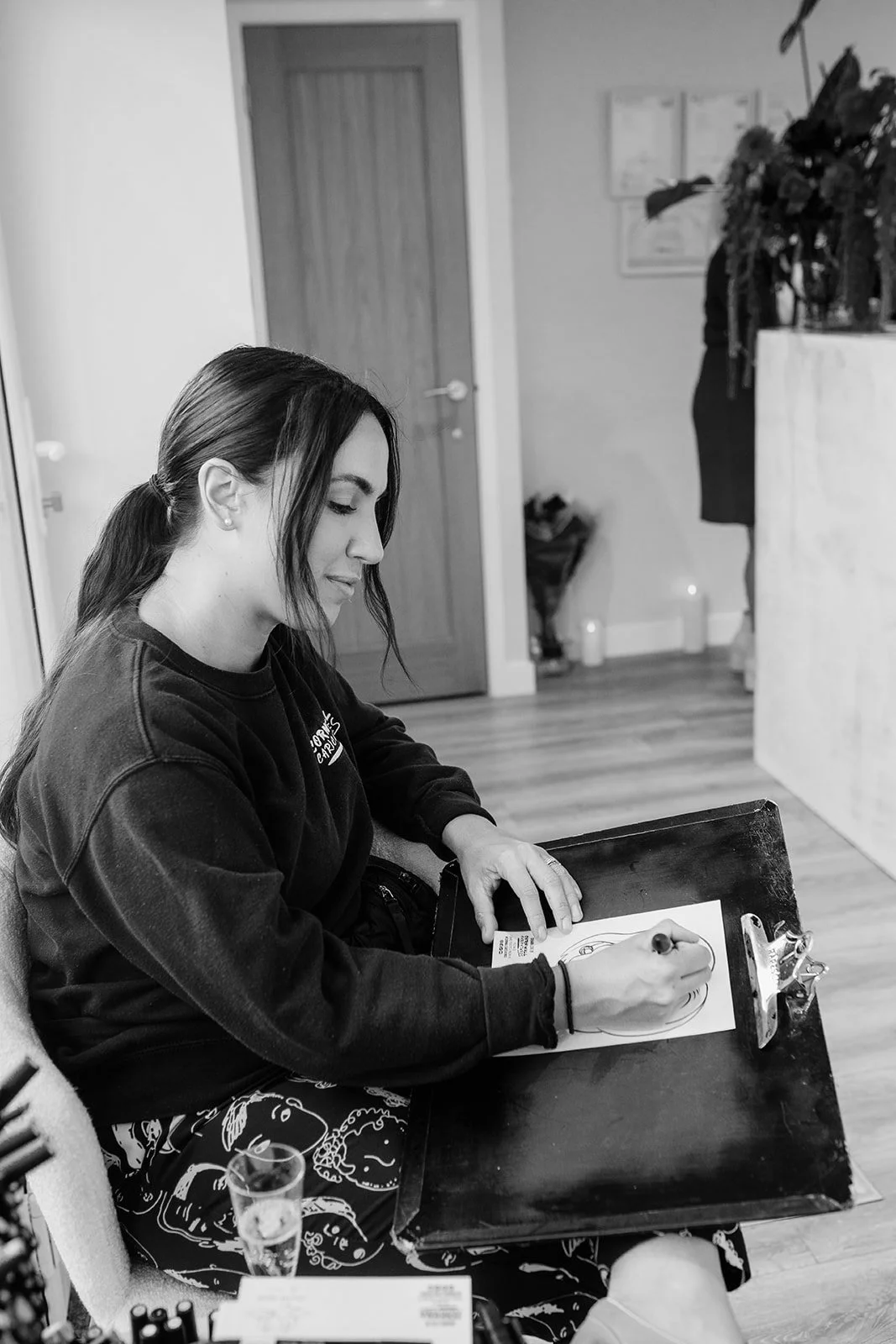 A woman with dark hair tied in a ponytail sitting at a desk, drawing on a piece of paper with a marker, in a room with a closed door and plants in the background.
