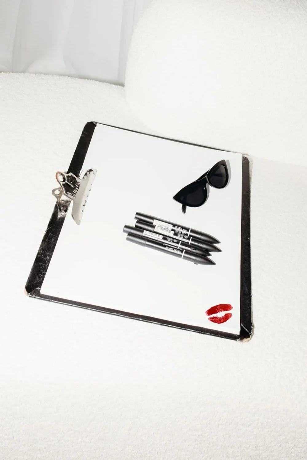 A rectangular mirror with a black frame resting on a white surface, reflecting a pair of black sunglasses, four black pens, and red lipstick kiss mark.